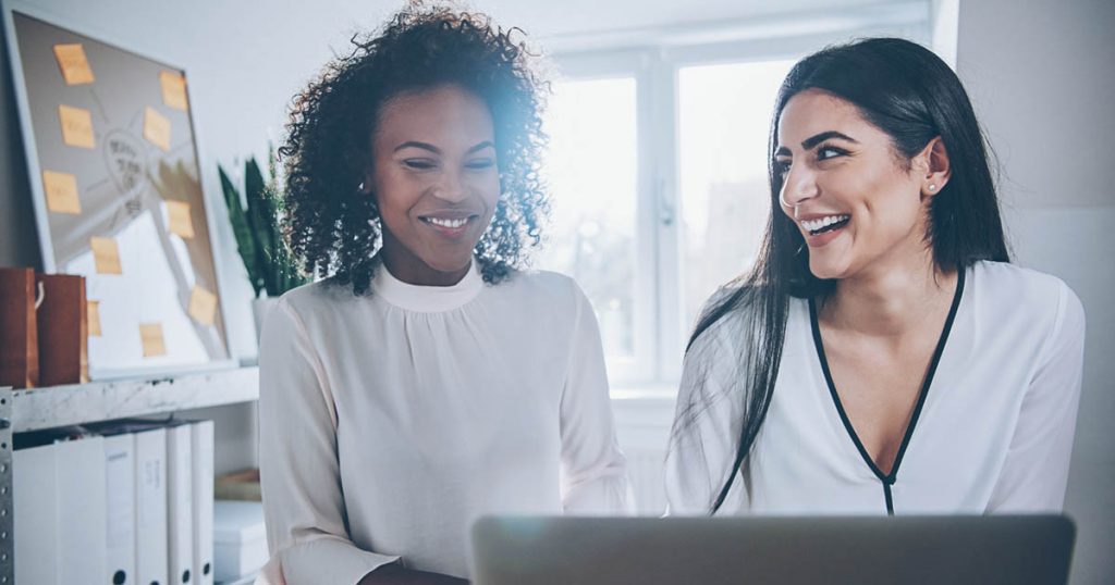 Two vivacious young businesswomen working together in a small business sitting laughing at something they have read on their laptop computer